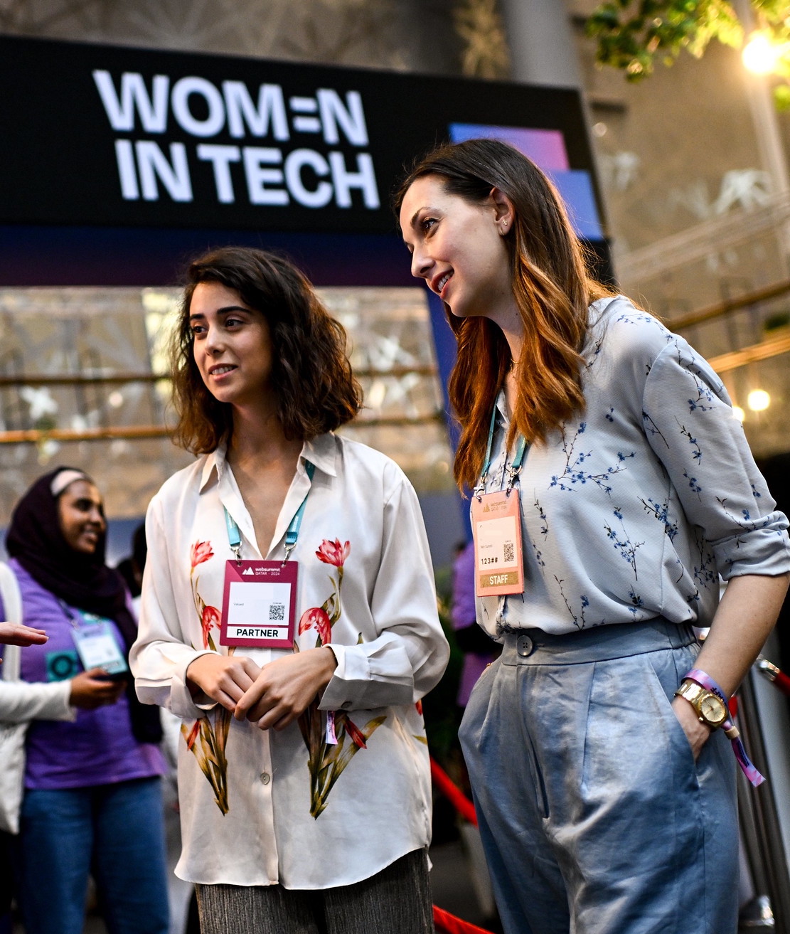 A photograph of two people at Web Summit Qatar, standing at the Women in Tech Lounge. They appear to be looking at another person that is not in frame.