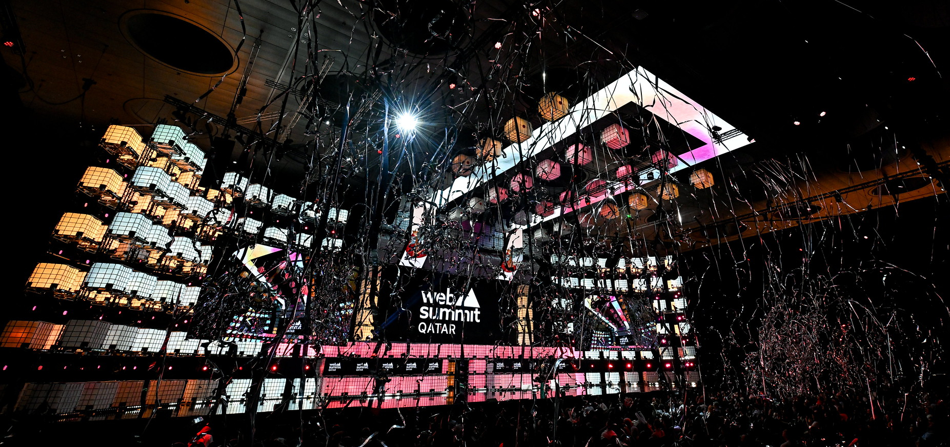 Streamers fall from an industrial ceiling above a large stage and crowded audience, viewed from the side. On the stage, a large screen shows the Web Summit Qatar logo. This is the closing ceremony of Web Summit Qatar.