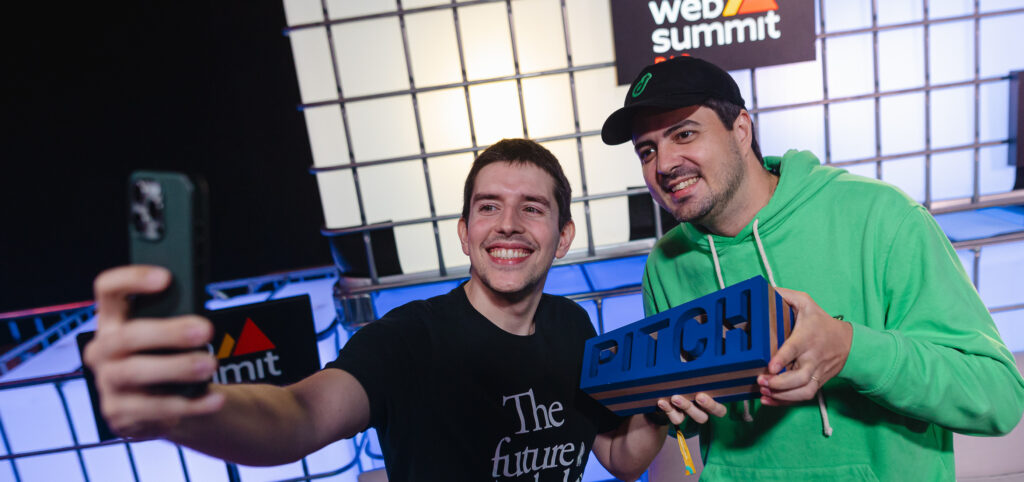 Guilherme Rodrigues and Rafael Crespo, co-founders of deco.cx, smile as they take a selfie of themselves holding the PITCH trophy on stage at Web Summit Rio.