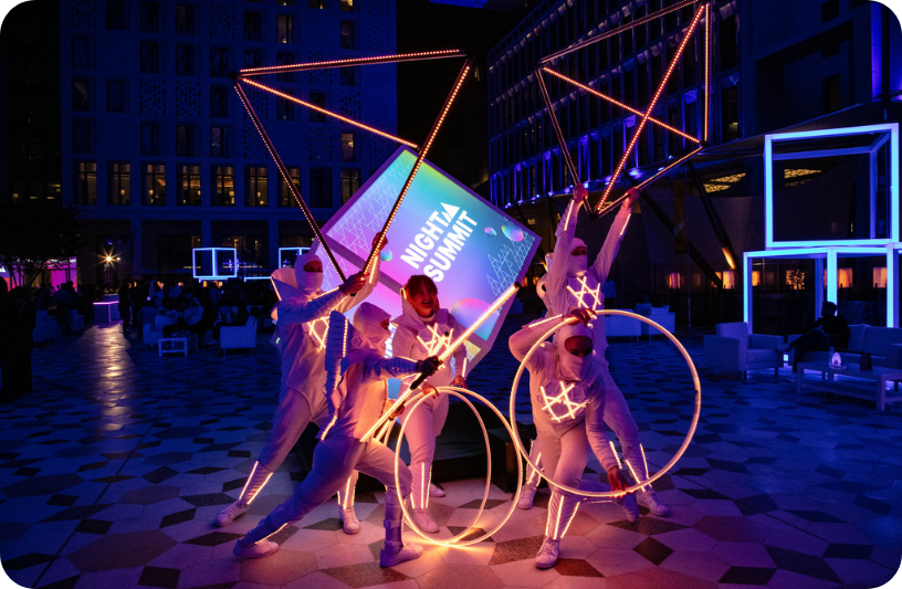 A photograph of Night Summit at Web Summit Qatar. There are five people dressed in morphsuits and they are holding triangular 3D objects. The objects are lit up. In the middle, there is a lit-up cube with the Night Summit logo.